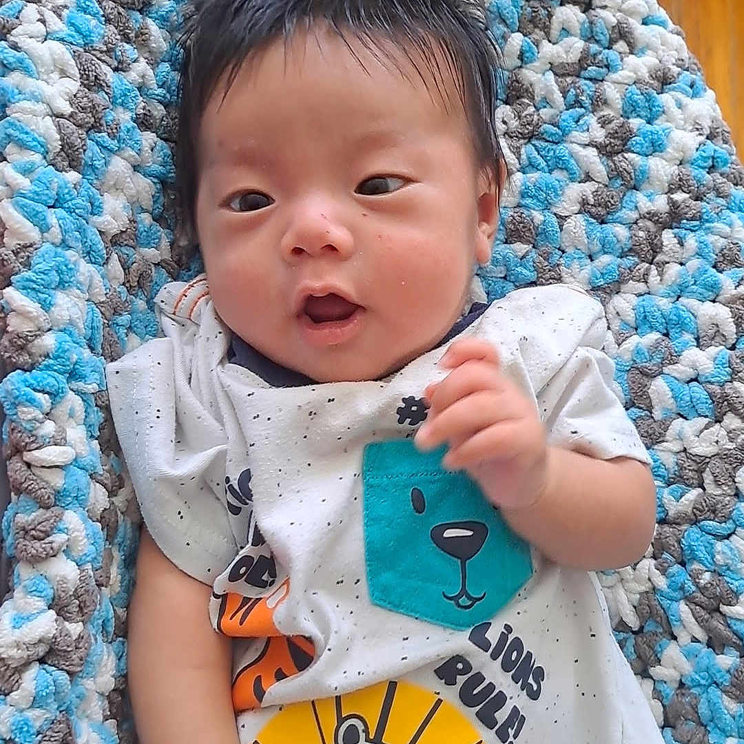 Khup Sang is registered to the contest to win money with this photo: baby, infant, child, tshirt, lion_graphic, blanket, knitted_blanket, blue, white, gray, wooden_floor, rug, indoor, cute, relaxed, expression, hair, hand, diaper, person