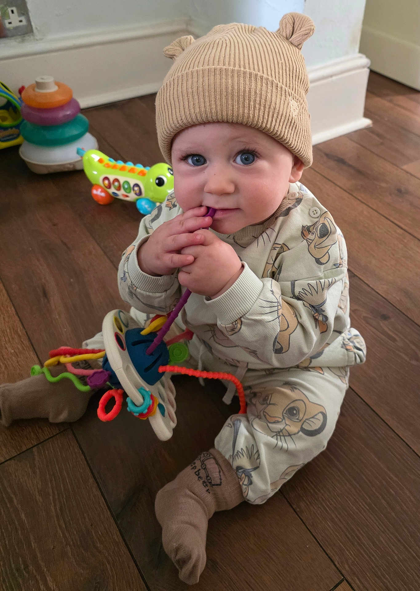 Carson is registered to the contest to win money with this photo: baby, child, hat, beige, ears, toy, colorful, floor, wooden_floor, sitting, clothing, cute, indoor, playful, infant, person, blue_eyes, long_sleeves, socks, smiling