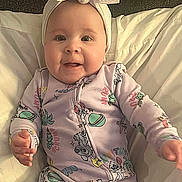 Briar is registered to the contest to win money with this photo: baby, bed, bow, cartoon_print, child, closeup, clothing, cute, face, happy, headband, indoors, infant, lying_down, onesie, person, pillow, portrait, small_hands, smiling