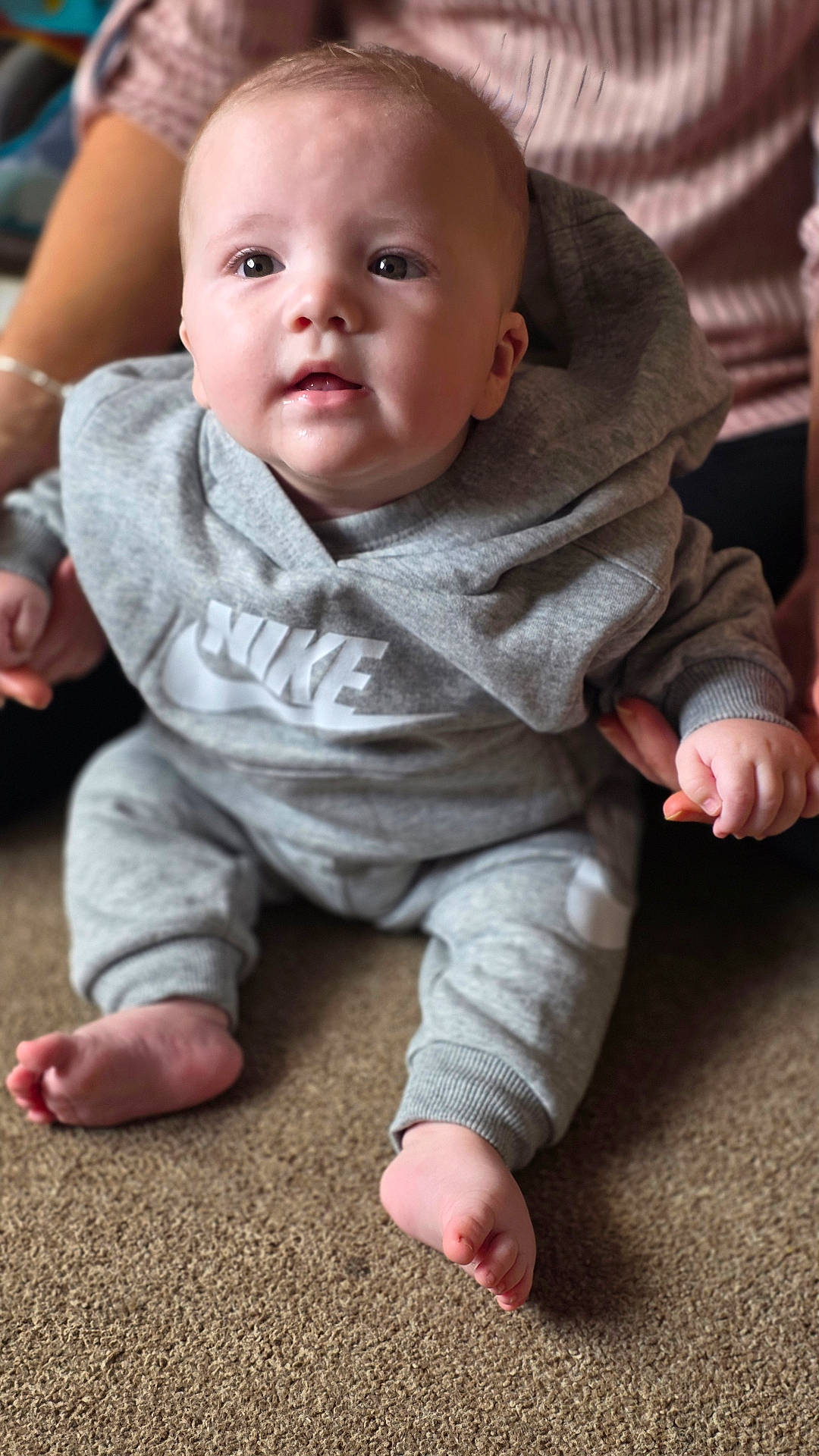 Abel joined the competition — help win amazing prizes! adult_hand, baby, barefoot, carpet, casual_wear, child, cute, grey_clothing, hoodie, indoor, infant, looking_up, nike, person, pink_sweater, portrait, sitting, soft_lighting, support, young_child