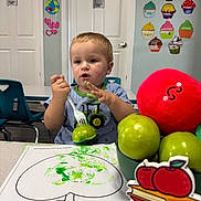 David joined the competition — help win amazing prizes! child, toddler, boy, portrait, sitting, classroom, table, fork, apple, fruit, red_ball, painting, art_project, green_paint, coloring, chair, doors, cupcake_decorations, indoors, smiling
