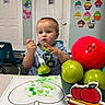 child, toddler, boy, portrait, sitting, classroom, table, fork, apple, fruit, red_ball, painting, art_project, green_paint, coloring, chair, doors, cupcake_decorations, indoors, smiling