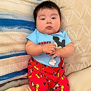 Erick is registered to the contest to win money with this photo: baby, infant, child, onesie, mickey_mouse, red_pants, sneakers, bracelet, pillow, bedding, headboard, couch, sitting, portrait, chubby_cheeks, dark_hair, cute, indoor, relaxed, clothing