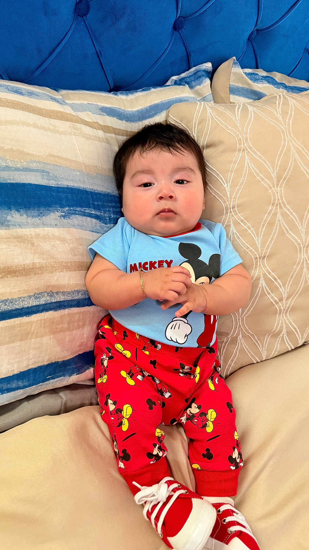 Erick is registered to the contest to win money with this photo: baby, infant, child, onesie, mickey_mouse, red_pants, sneakers, bracelet, pillow, bedding, headboard, couch, sitting, portrait, chubby_cheeks, dark_hair, cute, indoor, relaxed, clothing