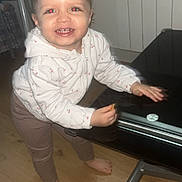 Inaya a rejoint le concours — aidez-le/la à gagner de superbes lots ! toddler, child, smiling, hoodie, barefoot, table, wooden_floor, indoor, red_eye, pants, person, standing, furniture, happy, cute, home, young_child, casual_clothing, face, hands