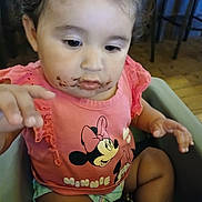 Skyelenna is registered to the contest to win money with this photo: toddler, child, smile, pink_shirt, minnie_mouse, chocolate, messy, high_chair, indoor, curly_hair, baby, hand, face, clothing, person, cute, sitting, expression, wood_floor, furniture