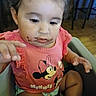 toddler, child, smile, pink_shirt, minnie_mouse, chocolate, messy, high_chair, indoor, curly_hair, baby, hand, face, clothing, person, cute, sitting, expression, wood_floor, furniture