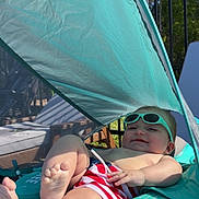 Liam is registered to the contest to win money with this photo: baby, infant, child, sunglasses, pool_float, sunshade, canopy, outdoor, summer, swim_trunks, striped_clothing, feet, legs, smile, reclining, turquoise, safety_device, fence, trees, relaxation