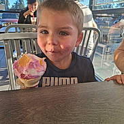 Mason is registered to the contest to win money with this photo: child, boy, ice_cream, cone, dessert, smiling, eating, food, cafe, restaurant, outdoor, sunlight, table, chair, window, blinds, background_people, portrait, puma_shirt, happy