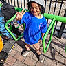 child, boy, hat, bucket_hat, blue_tshirt, peace_sign, smile, sneakers, pants, outdoor, pavement, metal_railings, fence, scooter, person, sunlight, shadow, playground, urban, casual_clothing