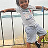 black_shoes, blue_sunglasses, boy, casual_clothing, child, cloudy_sky, curly_hair, daylight, gray_tshirt, greenery, happy, metal_railing, nature, outdoor, playful_pose, river, scenic_background, shorts, smiling, water