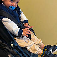 Shahine participe au concours pour gagner de l'argent avec cette photo : toddler, child, pacifier, stroller, sneakers, blue_eyes, jacket, pants, socks, indoor, seatbelt, curious, person, footwear, casual_clothing, young_child, baby_stroller, black_shoes, beige_background, seated