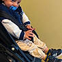 toddler, child, pacifier, stroller, sneakers, blue_eyes, jacket, pants, socks, indoor, seatbelt, curious, person, footwear, casual_clothing, young_child, baby_stroller, black_shoes, beige_background, seated