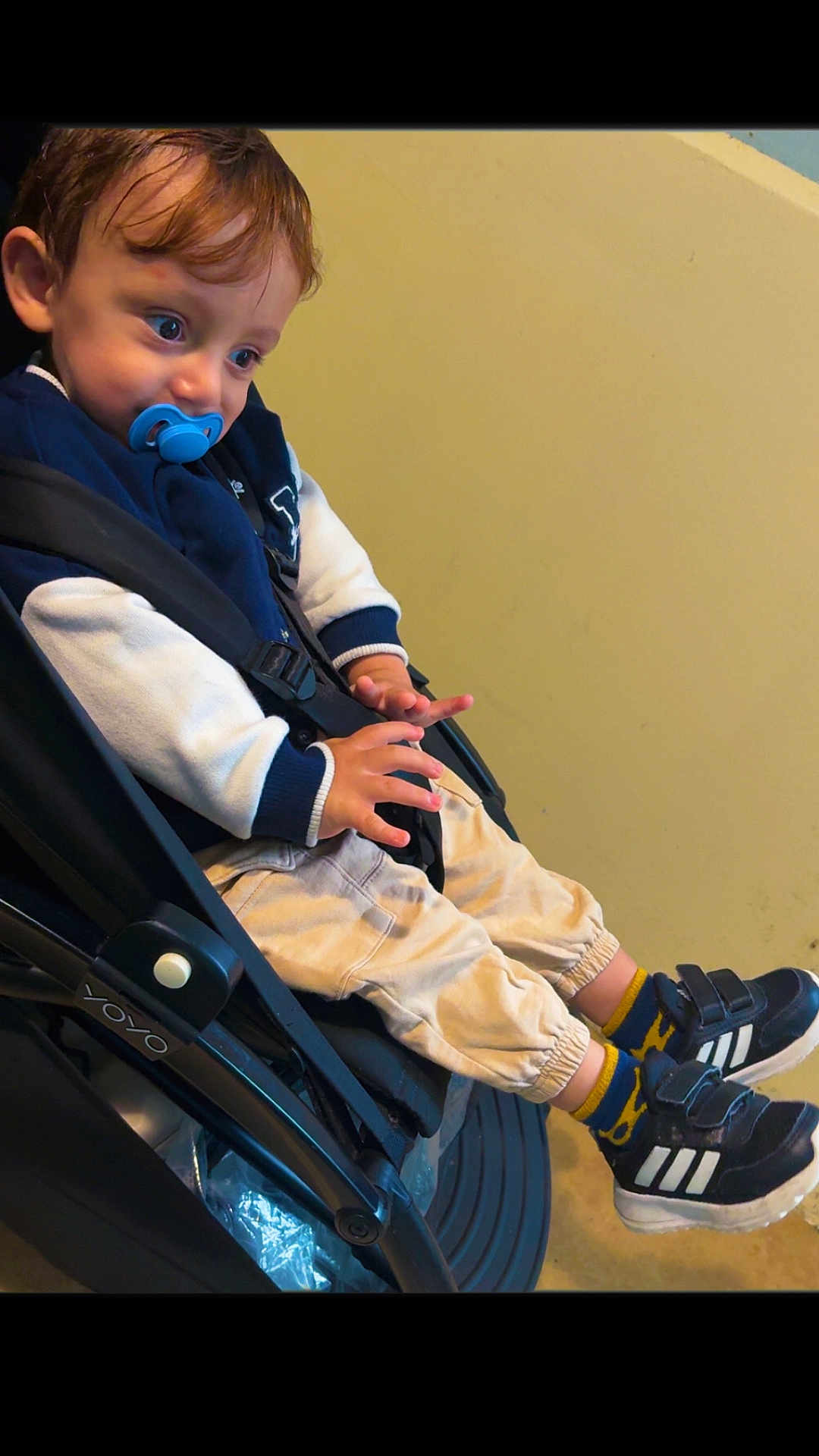 Shahine participe au concours pour gagner de l'argent avec cette photo : toddler, child, pacifier, stroller, sneakers, blue_eyes, jacket, pants, socks, indoor, seatbelt, curious, person, footwear, casual_clothing, young_child, baby_stroller, black_shoes, beige_background, seated