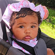 Elina is registered to the contest to win money with this photo: baby, child, stroller, hat, bonnet, curly_hair, outdoor, greenery, sunlight, cute, infant, face, portrait, seatbelt, pink, white, skin, expression, young_child, closeup