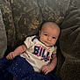 Maribel is registered to the contest to win money with this photo: baby, infant, child, person, couch, clothing, buffalo_bills, blue_skirt, patterned_cushion, smile, face, head, relaxing, indoor, furniture, text_on_clothing, cute, young, sitting, soft_fabric