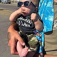 Kason is registered to the contest to win money with this photo: adult, baby, blue_towel, camouflage, casual, child, clothing, foot, hand, holding, outdoor, pacifier, parking_lot, person, shopping_cart, street, sunglasses, sunny_day, tattoo, walmart