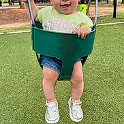 Amy joined the competition — help win amazing prizes! toddler, child, swing, playground, grass, sneakers, hair_clip, green_swing, smiling, happy, outdoor, park, shorts, tshirt, daylight, trees, people, background, metal_chains, fun
