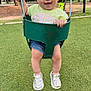 toddler, child, swing, playground, grass, sneakers, hair_clip, green_swing, smiling, happy, outdoor, park, shorts, tshirt, daylight, trees, people, background, metal_chains, fun