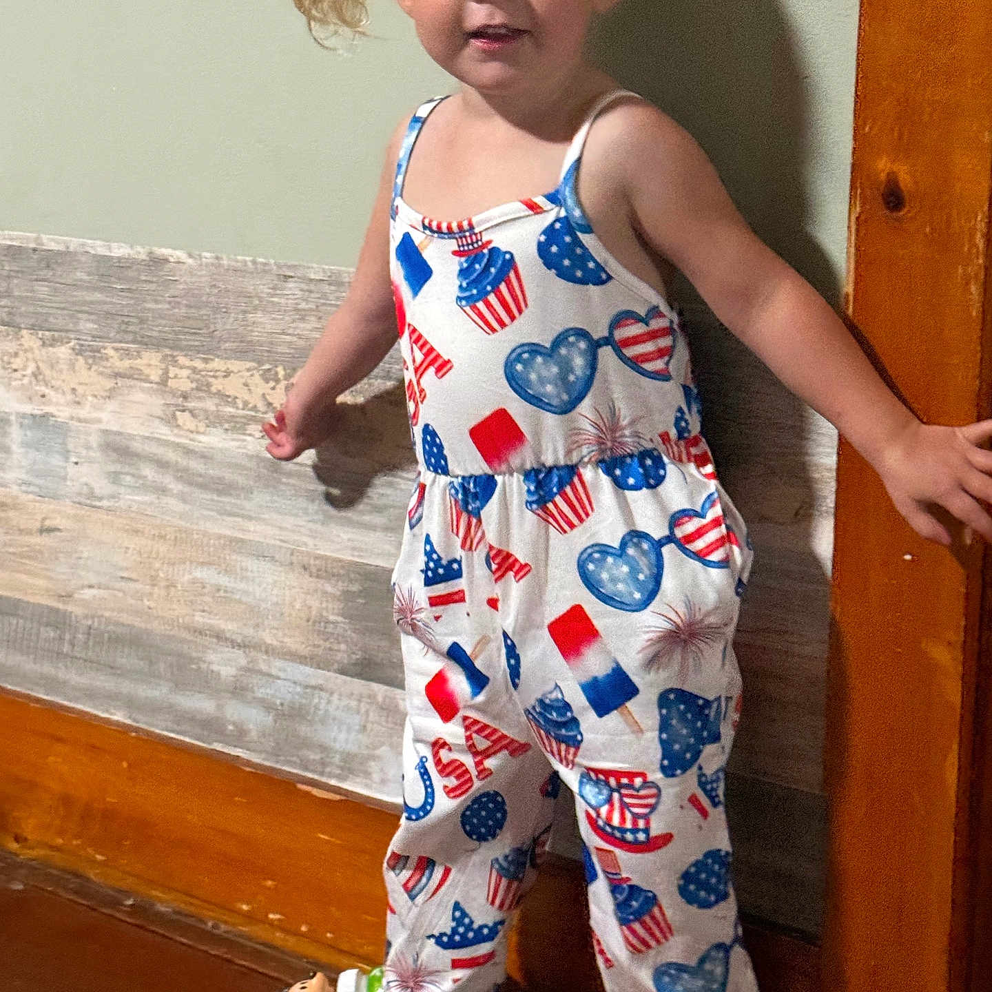 Olivia is registered to the contest to win money with this photo: barefoot, blonde_hair, casual, child, clothing, cute, home, indoor, jumpsuit, person, pigtails, playful, smiling, standing, toddler, toy_bottle, usa_theme, wooden_floor, wooden_wall, young_child
