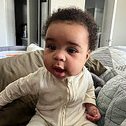 Claire Faith is registered to the contest to win money with this photo: baby, child, infant, onesie, toy, bed, pillow, blanket, indoor, curly_hair, open_mouth, sitting, cute, portrait, home, room, door, furniture, comfort, young