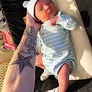 Jamison is registered to the contest to win money with this photo: baby, newborn, infant, child, tattoo, arm, blue_clothing, hat, onesie, socks, changing_pad, person, indoor, soft_lighting, cute, face, hand, skin, resting, portrait