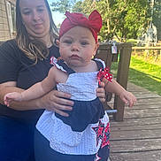 Sophia is registered to the contest to win money with this photo: baby, child, woman, outdoor, deck, wooden_deck, trees, sky, headband, red_bow, dress, star_pattern, holding, person, smile, nature, sunny, casual_clothing, portrait, family