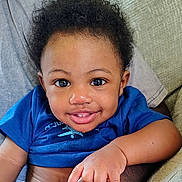Logann is registered to the contest to win money with this photo: baby, child, smiling, portrait, close_up, curly_hair, blue_shirt, sitting, indoor, couch, blanket, face, eyes, hands, arm, wristband, happy, cozy, infant, photography