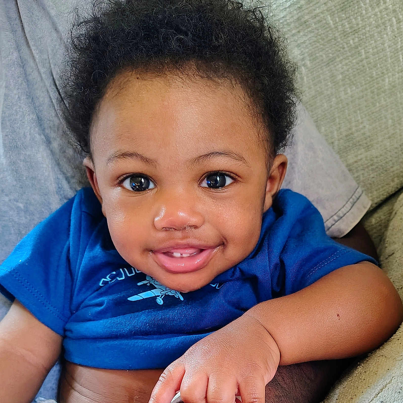 Logann is registered to the contest to win money with this photo: baby, child, smiling, portrait, close_up, curly_hair, blue_shirt, sitting, indoor, couch, blanket, face, eyes, hands, arm, wristband, happy, cozy, infant, photography