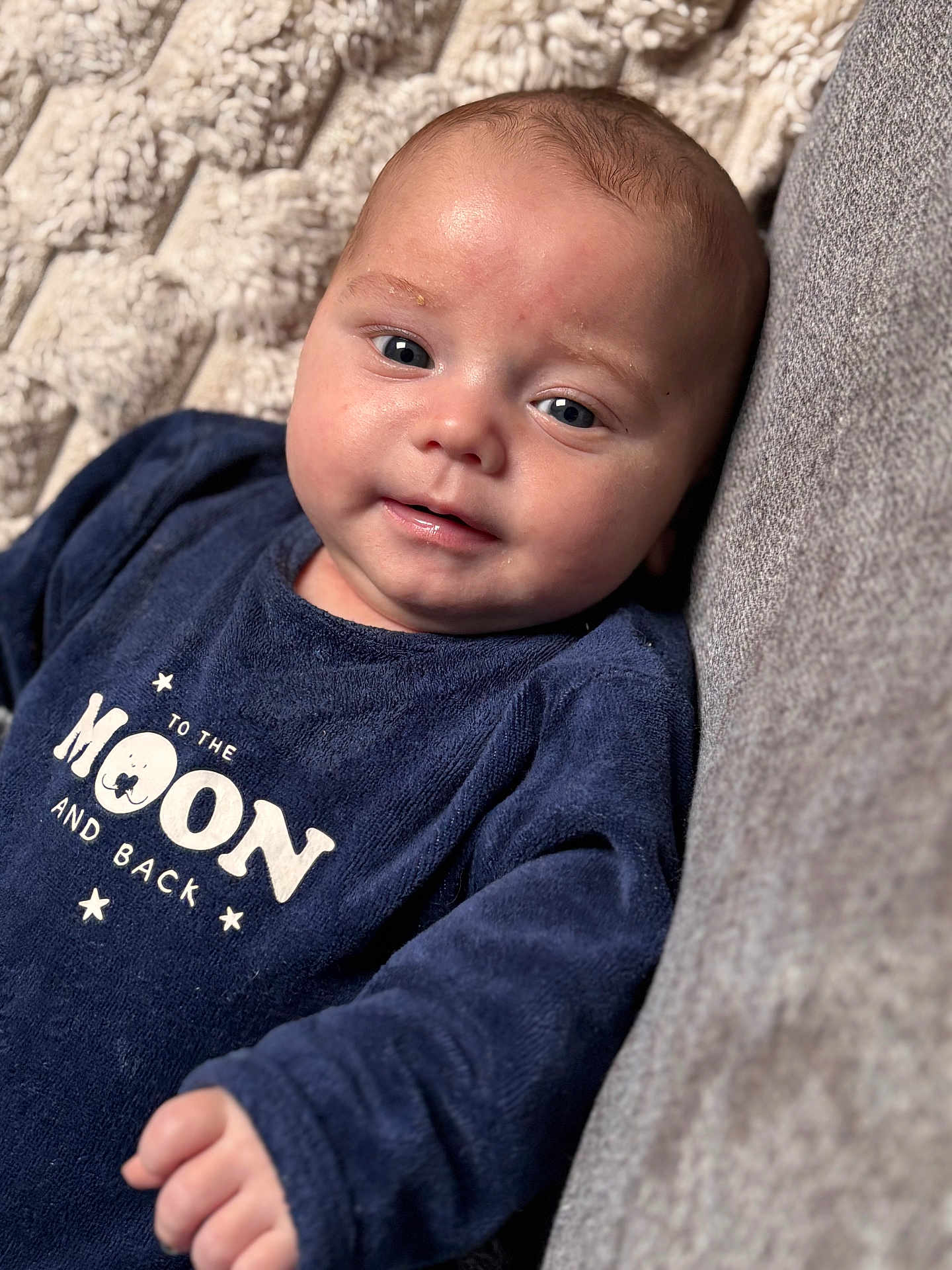 Lyzio participe au concours pour gagner de l'argent avec cette photo : baby, infant, face, eyes, smile, clothing, blue_outfit, text_on_clothing, blanket, couch, texture, indoor, portrait, young_child, skin, head, hand, cute, lying_down, cozy