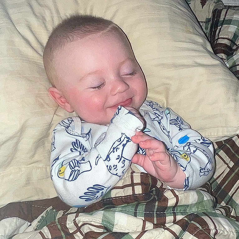 Forrestt is registered to the contest to win money with this photo: baby, child, sleeping, smiling, pajamas, blanket, bed, pillow, cozy, face, infant, cute, indoor, resting, comfort, patterned, plaid, hands, peaceful, soft_light
