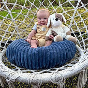 Chance is registered to the contest to win money with this photo: baby, child, outdoor, hammock, cushion, stuffed_animal, toy, grass, flowers, smiling, happy, overalls, plush, seat, nature, greenery, sunlight, person, cute, relaxing