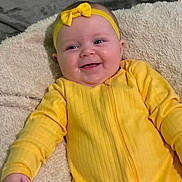 Aries is registered to the contest to win money with this photo: baby, infant, yellow_outfit, headband, bow, smile, happy, blanket, soft_texture, lying_down, cute, child, indoor, portrait, face, small_hand, cheeks, eyes, clothing, warm_colors