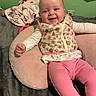baby, smiling, infant, pink_leggings, floral_vest, cushion, blanket, bed, green_wall, happy, person, indoor, cute, child, portrait, clothing, face, sitting, young_child, cozy