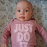 baby, infant, onesie, pink_clothing, crib, crib_sheet, patterned_sheet, face, eyes, tongue, child, cute, portrait, lying_down, indoors, baby_garment, slogan, expression, skin, head