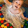 child, toddler, ball_pit, colorful_balls, yellow_ball, dress, daisies, smiling, happy, playful, indoor, cute, fun, person, baby, face, hair, skin, sitting, toy