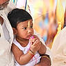 toddler, child, baby, portrait, sitting, indoors, celebration, family, adult, man, traditional_clothing, white_clothing, black_hair, facial_mark, bracelets, necklace, pink_toy, hands, colorful_background, candid