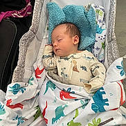 Zane joined the competition — help win amazing prizes! baby, infant, sleeping, bassinet, blanket, dinosaur_pattern, blue_plush, nursery, portrait, cozy, indoors, lying_down, peaceful, newborn, clothing, bedding, crib, face, resting, colorful