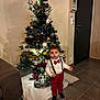 child, toddler, christmas_tree, holiday_decorations, red_bow_tie, suspenders, white_shirt, red_pants, black_shoes, indoor, living_room, couch, floor_tiles, ornaments, pine_cones, lights, festive, curious, holiday, decor