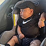 baby, car_seat, child, black_clothing, cap, bracelet, footwear, indoor, infant, looking_away, person, seat, sitting, side_view, sporty, window, young_child, cute, casual, relaxed