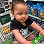 baby, child, shopping_cart, grocery_store, snacks, chips, toddler, clothing, black_shirt, bracelet, face, hand, food, aisle, retail, supermarket, person, expression, portrait, shopping