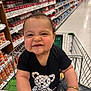 baby, child, shopping_cart, grocery_store, canned_goods, aisle, black_shirt, bear_graphic, bracelet, smiling, cute, face, person, holding, metal_cart, floor, retail, indoor, happy, toddler
