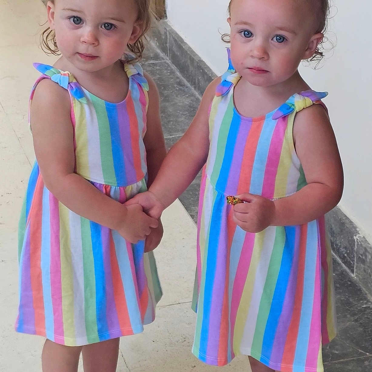 Charlotte And Rylei is registered to the contest to win money with this photo: blue_eyes, child, curious_expression, cute, holding_hands, indoor, person, pony_tail, portrait, purple_shoes, rainbow_dress, shoes, standing, striped_clothing, tiled_floor, toddler, twins, white_shoes, white_wall, young_children