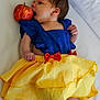 baby, infant, costume, dress, blue_top, yellow_skirt, red_bow, apple, fruit, lying_down, white_background, curious, hand, foot, face, head, skin, newborn, child, cute