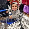toddler, shopping_cart, reindeer_antlers, smiling, child, store, clothing_rack, holiday_hat, indoor, person, happy, shopping, casual_clothing, retail, floor, footwear, adult, gray_jacket, blue_seat, festive