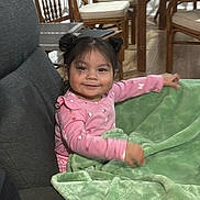 Elina is registered to the contest to win money with this photo: toddler, child, smiling, pink_clothing, blanket, green_blanket, indoor, furniture, couch, wooden_chairs, table, hair_buns, happy, person, home, floor, wood_floor, seated, cute, portrait