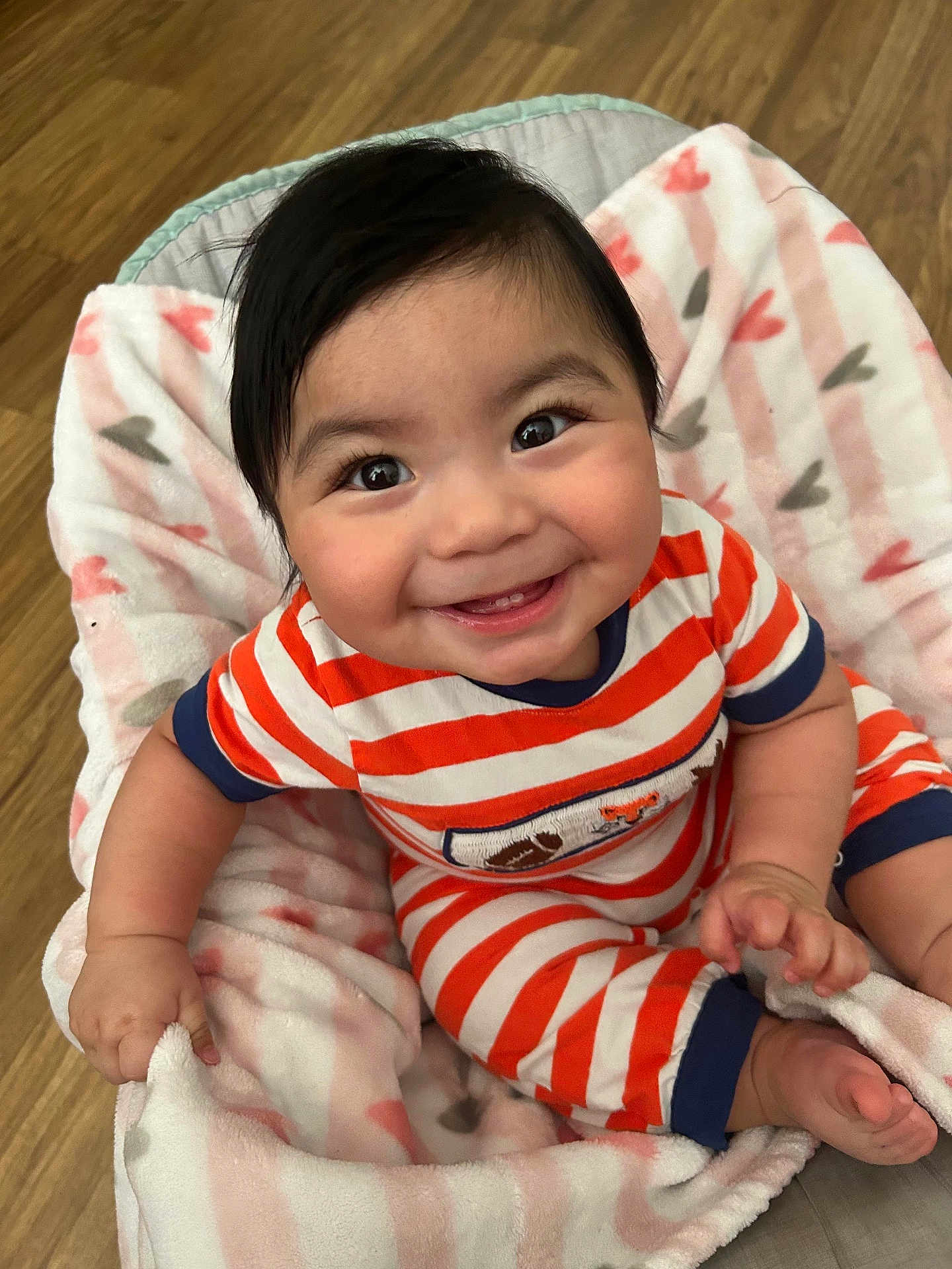 Elias is registered to the contest to win money with this photo: baby, smiling, child, striped_clothing, blanket, heart_pattern, happy, infant, cute, indoor, person, soft_texture, floor, wooden_floor, sitting, toes, hand, face, portrait, cozy