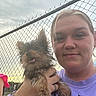 Princess is registered to the contest to win money with this photo: woman, dog, pet, outdoor, fence, chain_link_fence, sky, cloudy, smile, holding, small_dog, casual, portrait, happy, animal, person, fur, cute, daytime, watch