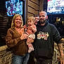 family, grandmother, toddler, teenager_peeking, bearded_man, tattoos, hoodie, holding_baby, indoor, restaurant, stone_wall, television, sports_broadcast, smiling, jeans, sneakers, window, group_photo, casual_clothing, eyeglasses_on_head