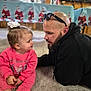 child, toddler, man, adult, beard, sunglasses, bow, pink_clothing, carpet, indoor, table, banner, santa_decor, interaction, portrait, face, gaze, earrings, cute, family_moment